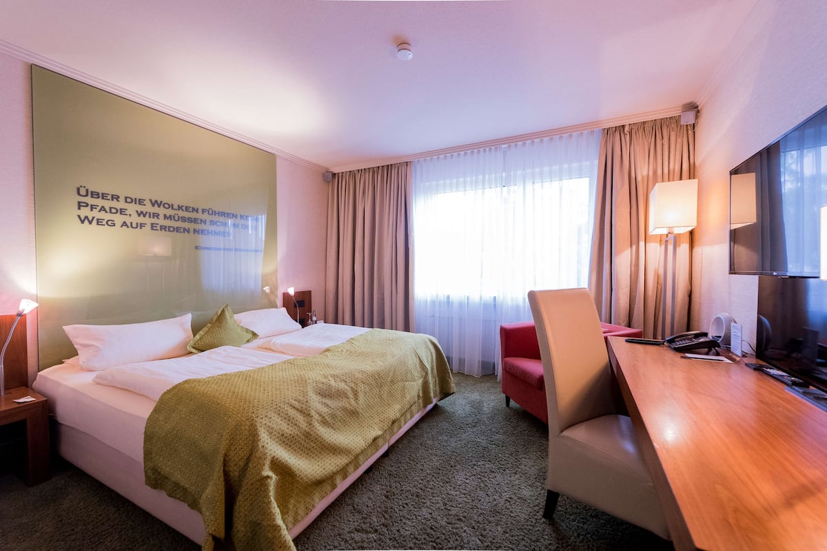 Ett hotellrum med en säng, ett skrivbord, en stol, en lampa och en stor spegel.