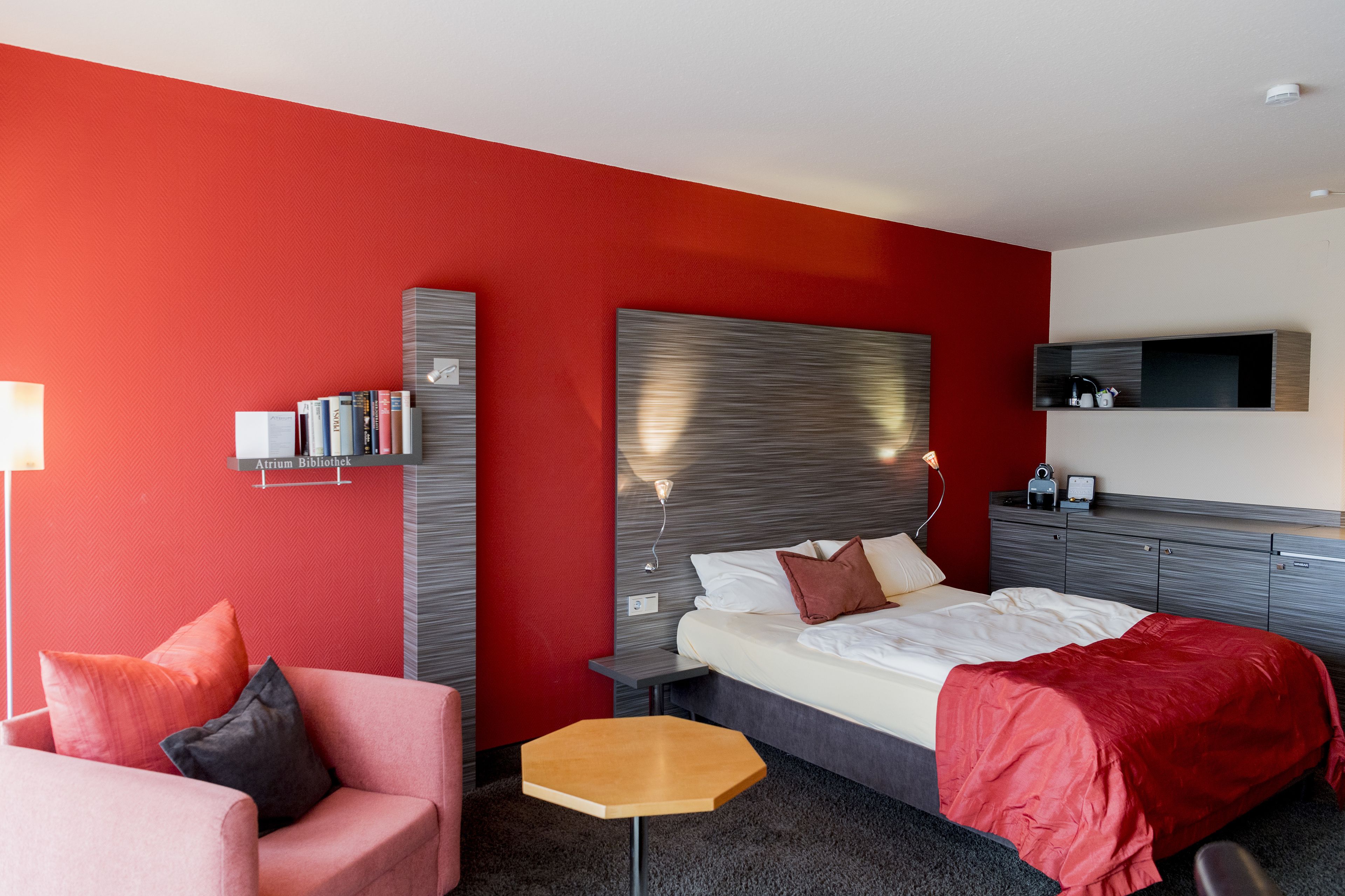 Una habitación de hotel moderna con una pared de acento roja, una cama grande con cabecero gris, una mesita de noche de madera y un sillón rosa, además de un mueble empotrado para la televisión.