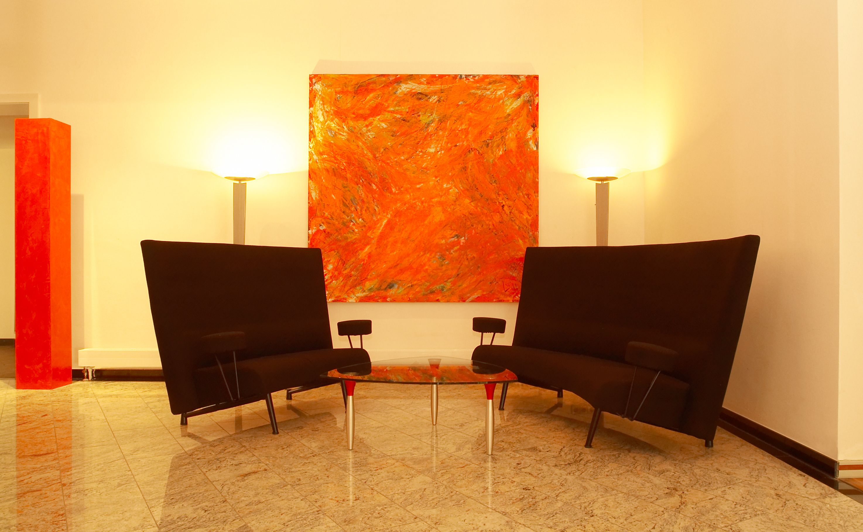 Un lobby moderno con dos sillones negros, una mesa de centro de vidrio y un cuadro abstracto grande de color naranja en la pared.