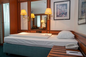 Una habitación de hotel con una cama, lámparas de noche, teléfono, un cuadro enmarco y un espejo.