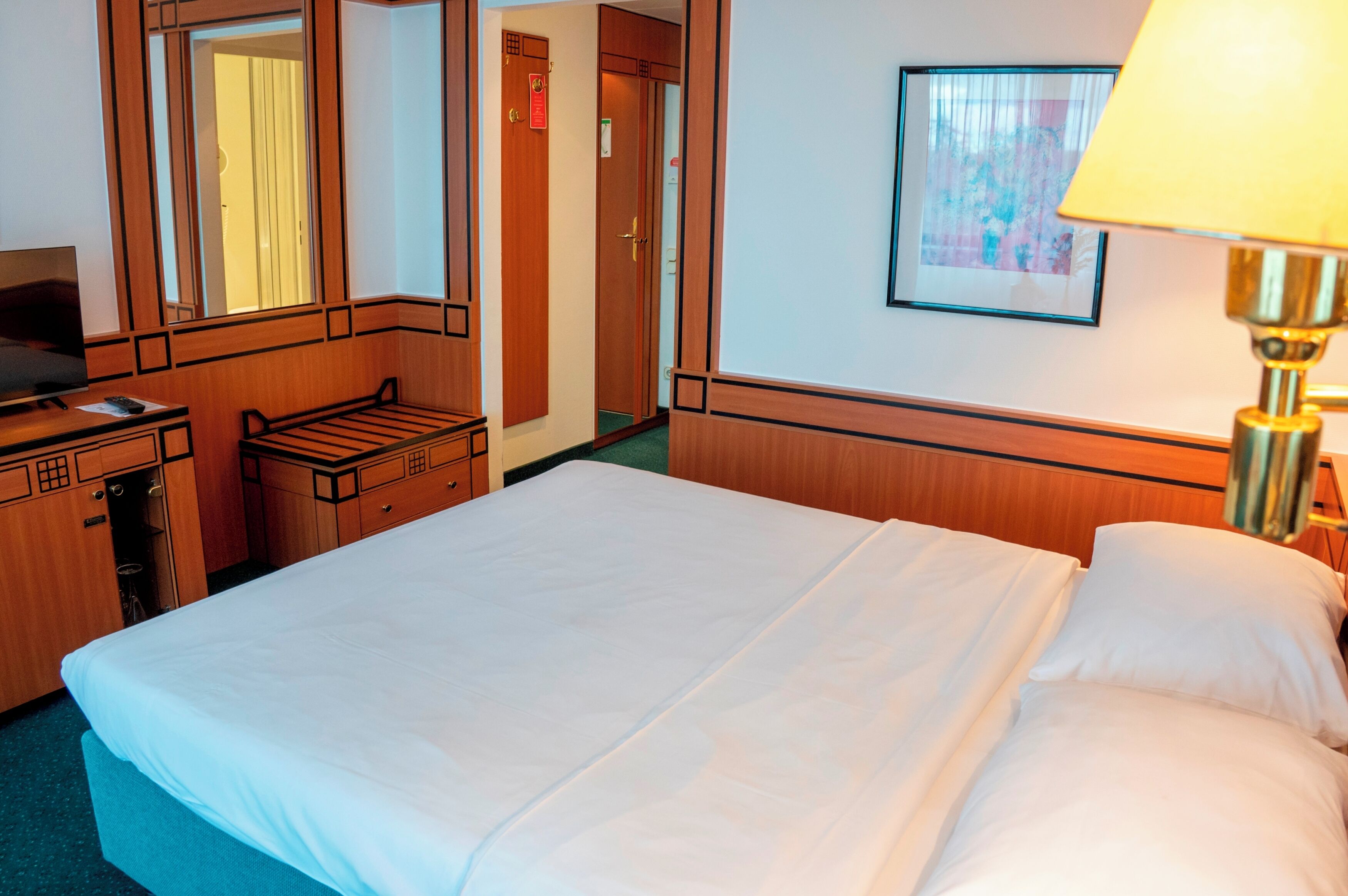 A hotel room with a bed, a desk with a computer, a lamp, and a framed picture on the wall.