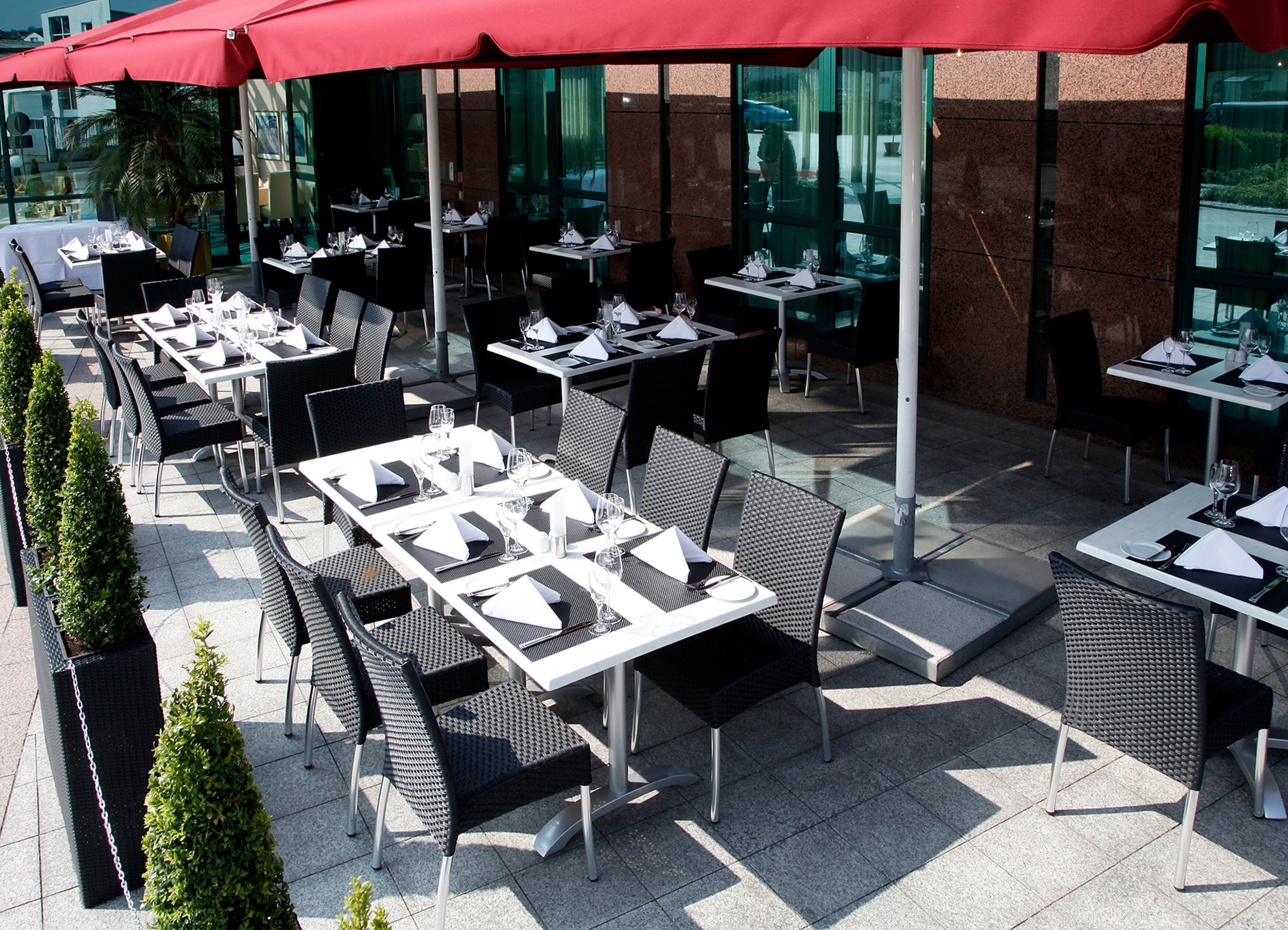 An outdoor dining area with tables and chairs, red umbrellas, and a modern building in the background.
