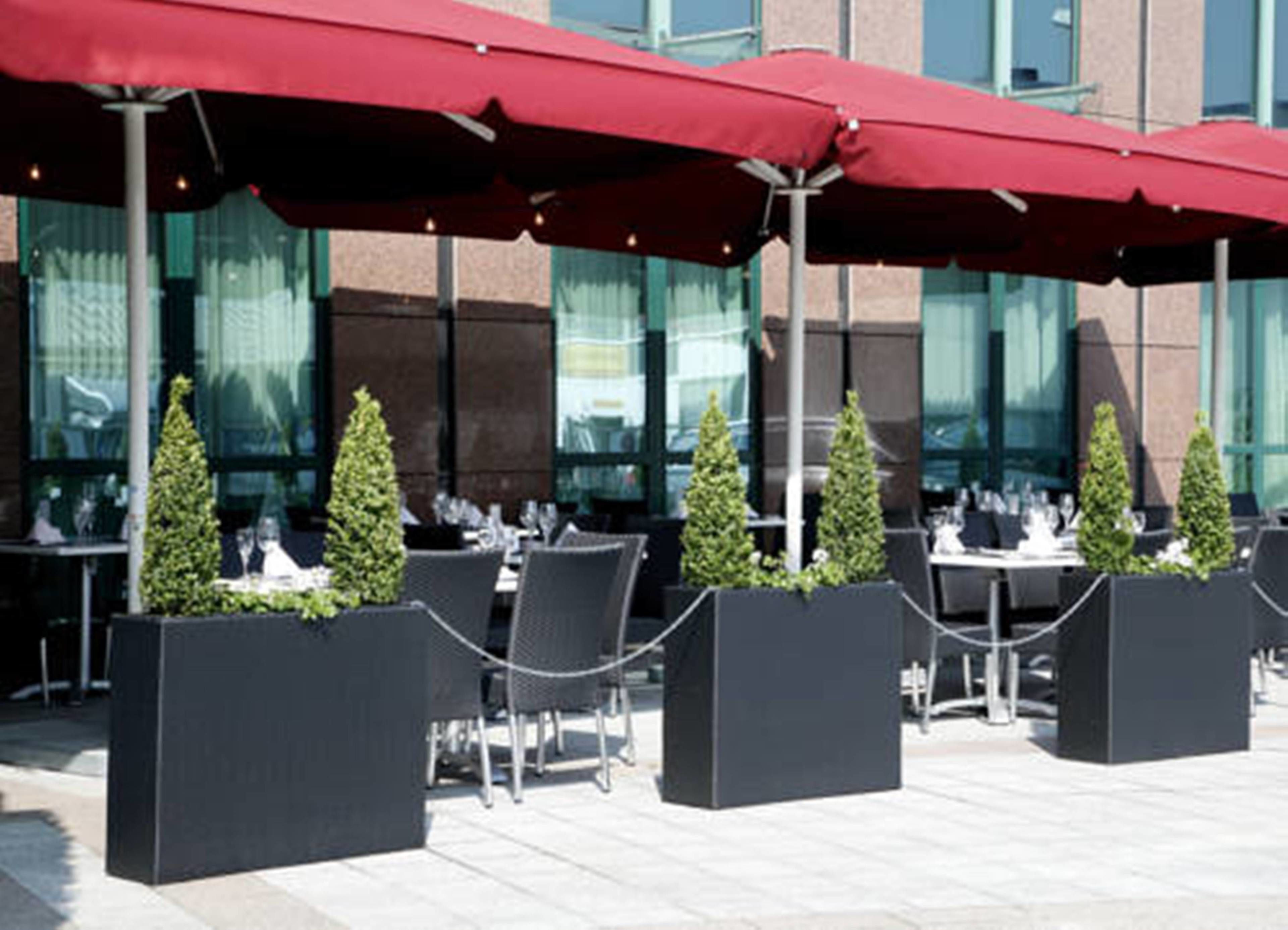 Un espace extérieur aménagé pour les repas, avec des tables et des chaises, des parasols rouges et de petites plantes en pot.