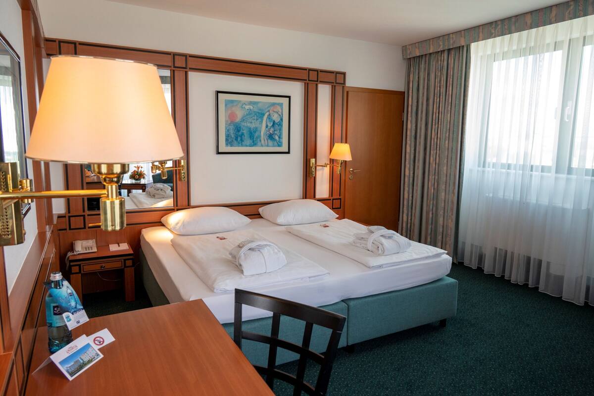 A hotel room with a bed, two bedside tables, a desk, a chair, a lamp, a painting, and a window with curtains.
