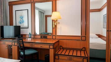 A hotel room with a wooden desk, a chair, a television, a bed, a bedside table, and a lamp.