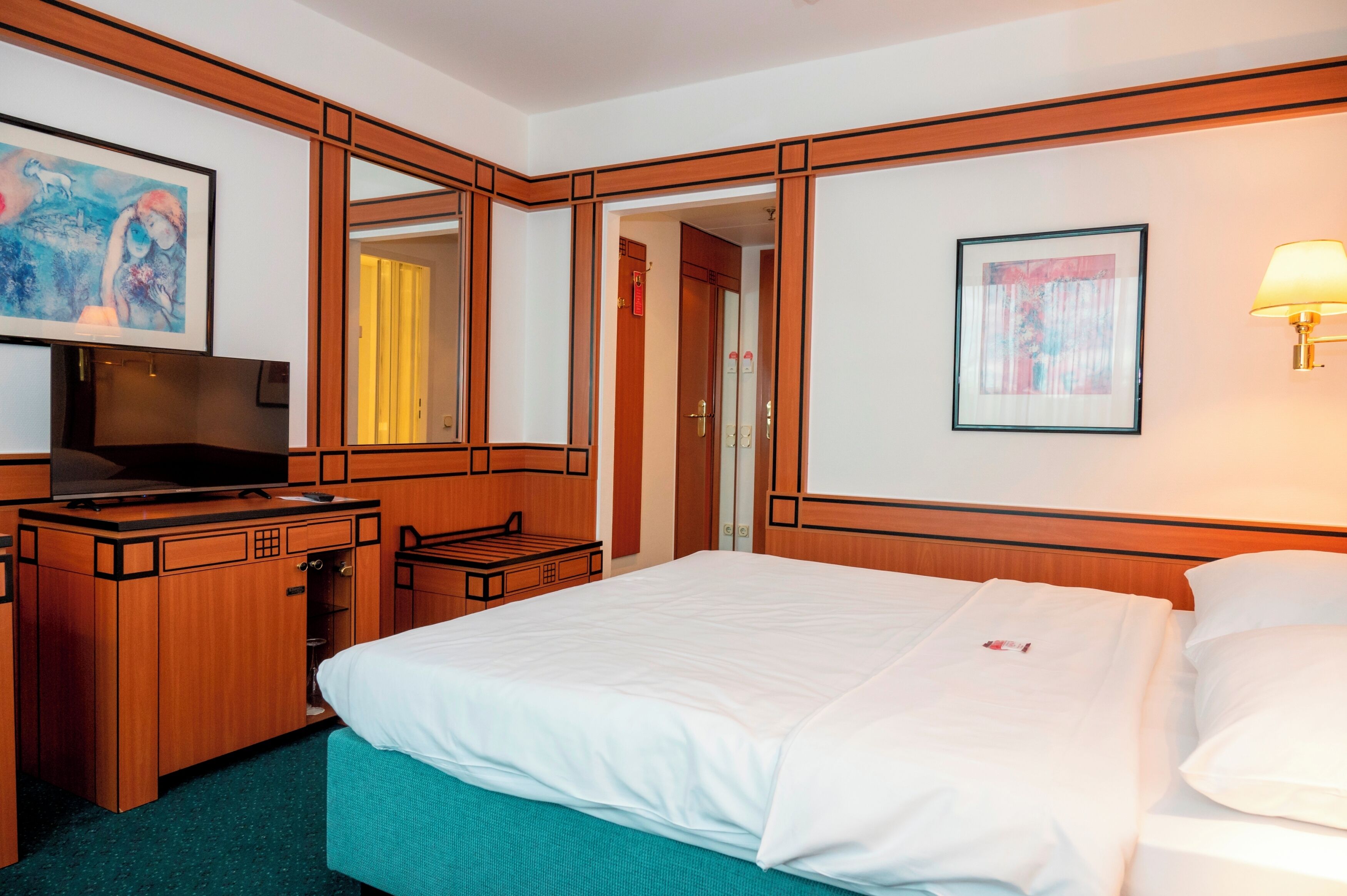 A hotel room with a bed, a desk with a television, a mirror, and artwork on the walls.