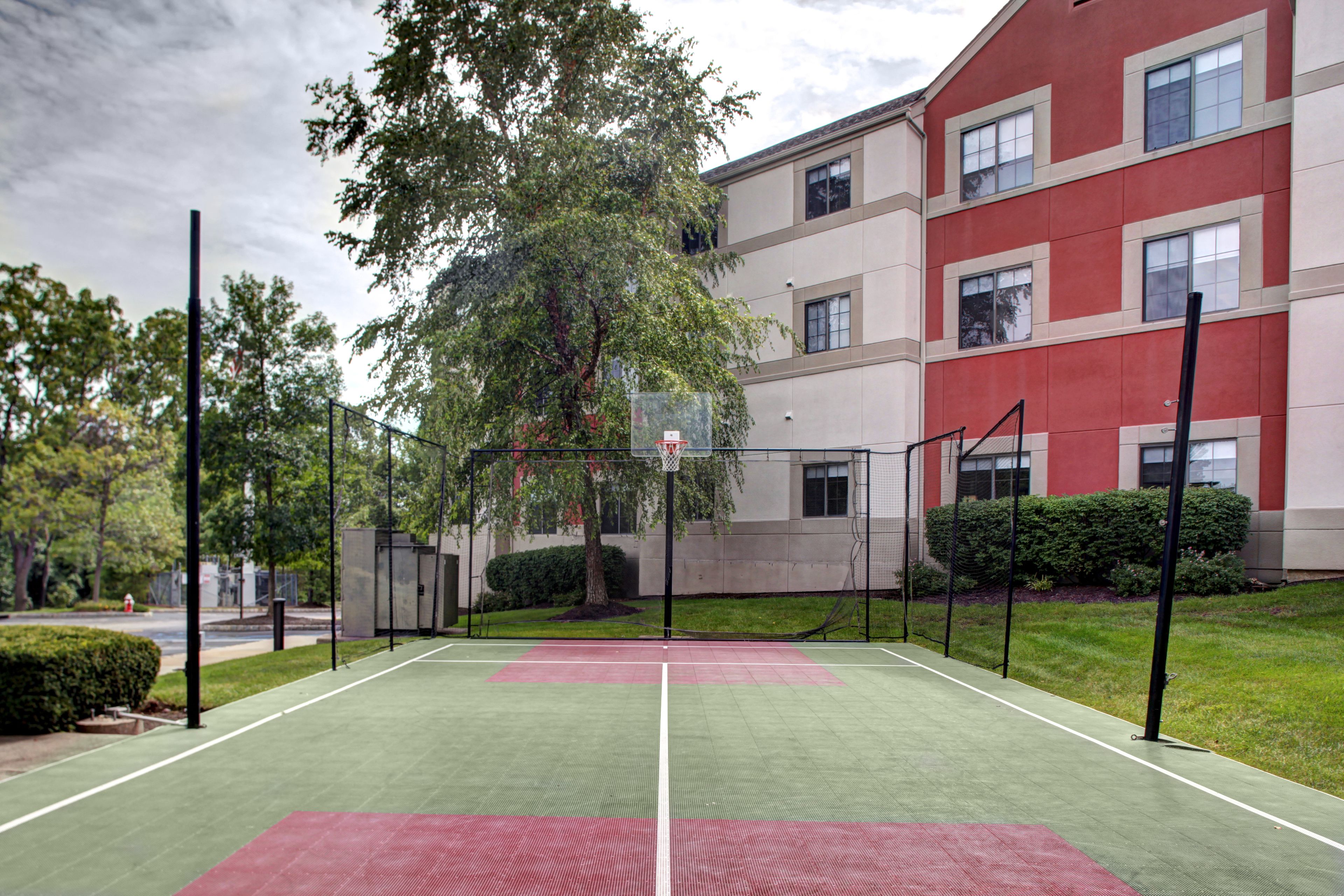 An outdoor tennis court with a net, surrounded by trees and a building with multiple windows.