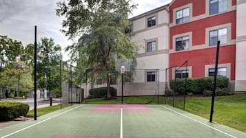An outdoor tennis court with a net, surrounded by trees and a building with multiple windows.