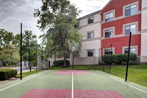 Cancha de tenis al aire libre con red, rodeada de árboles y un edificio con numerosas ventanas.
