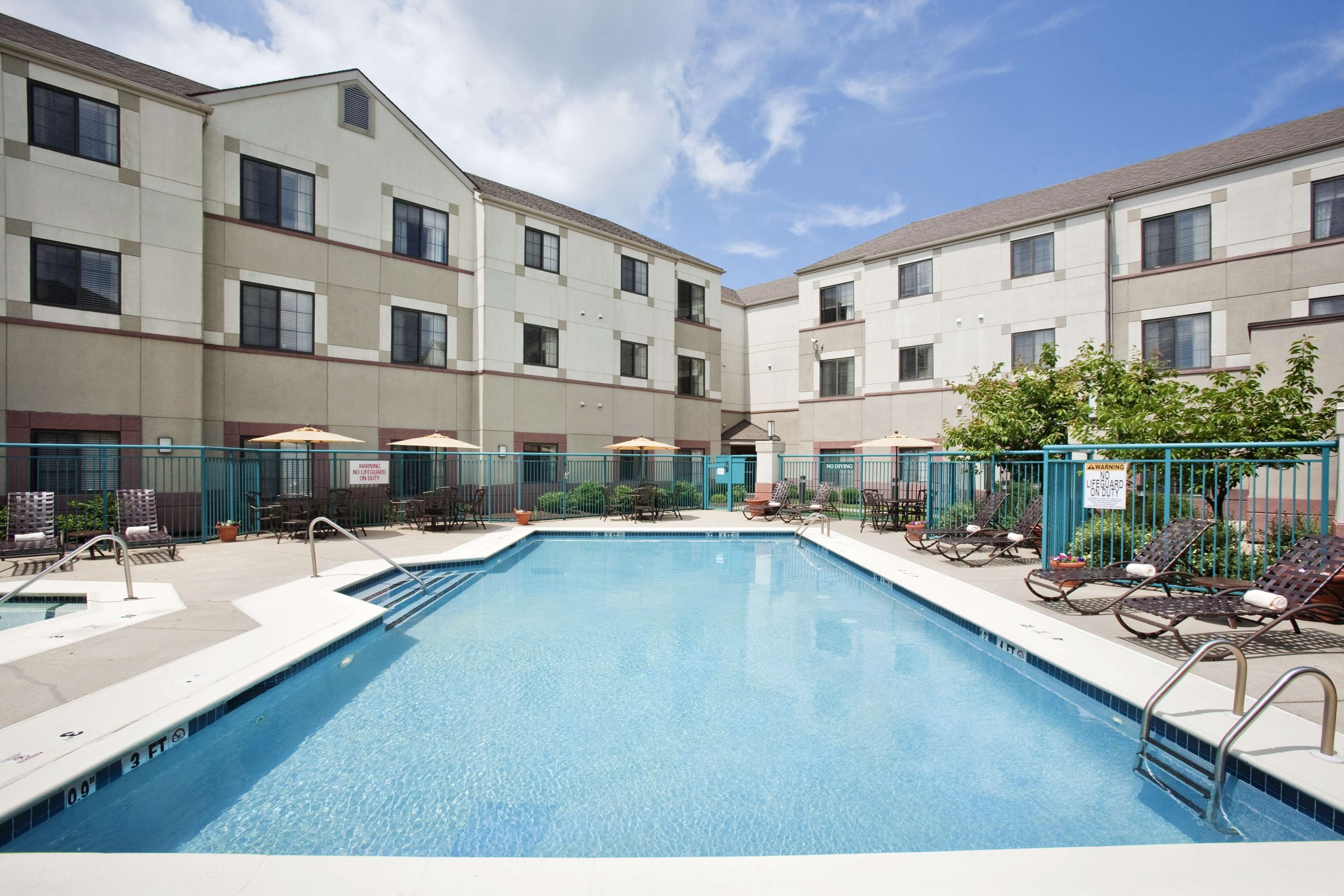 A swimming pool with lounge chairs and umbrellas, surrounded by a fence and a building with multiple windows.