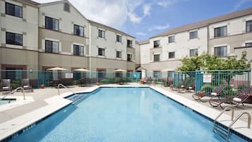 A swimming pool with lounge chairs and umbrellas, surrounded by a fence and a building with multiple windows.