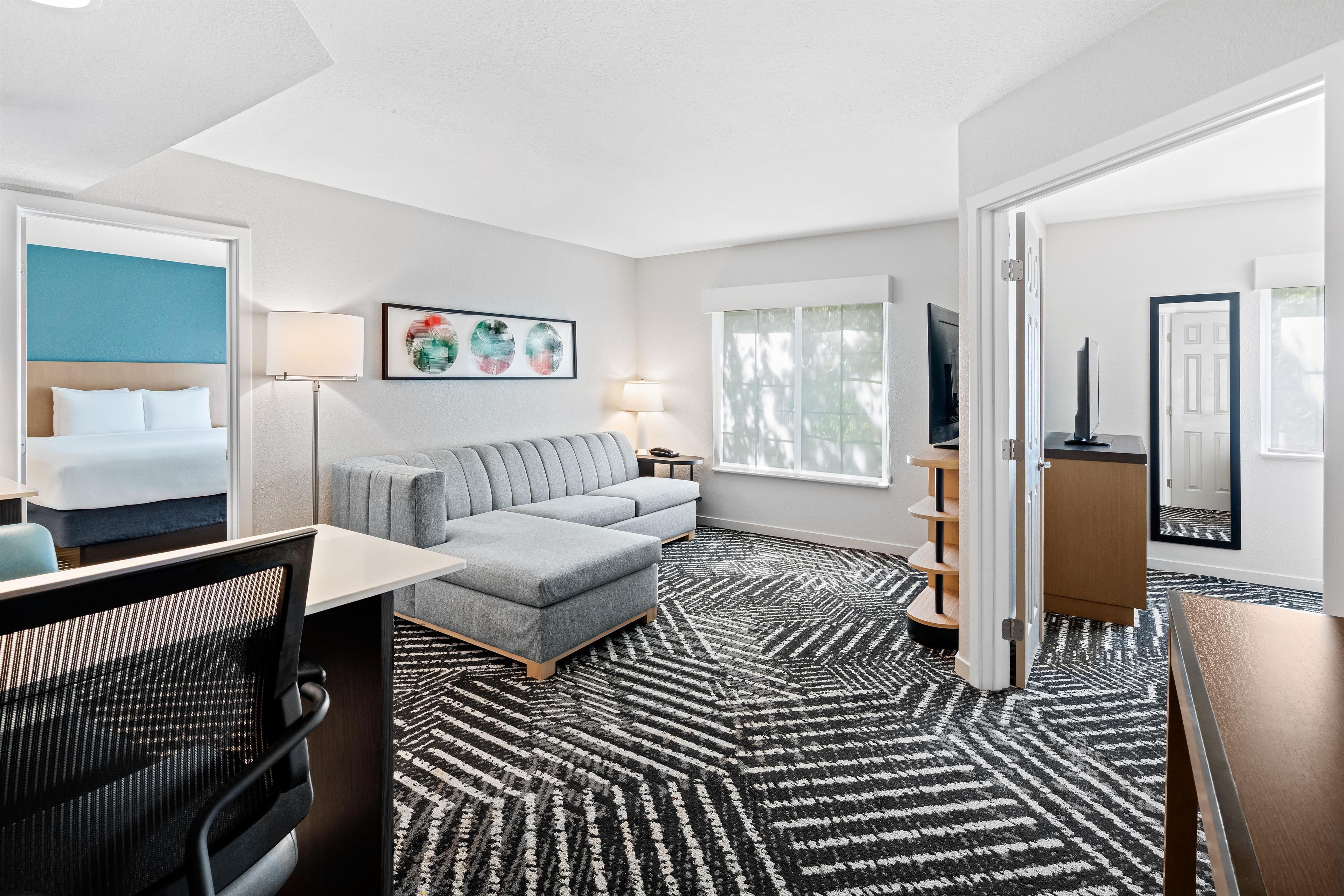 Habitación de hotel con cama, sofá, escritorio, silla, televisor, lámpara, estantería y ventana.