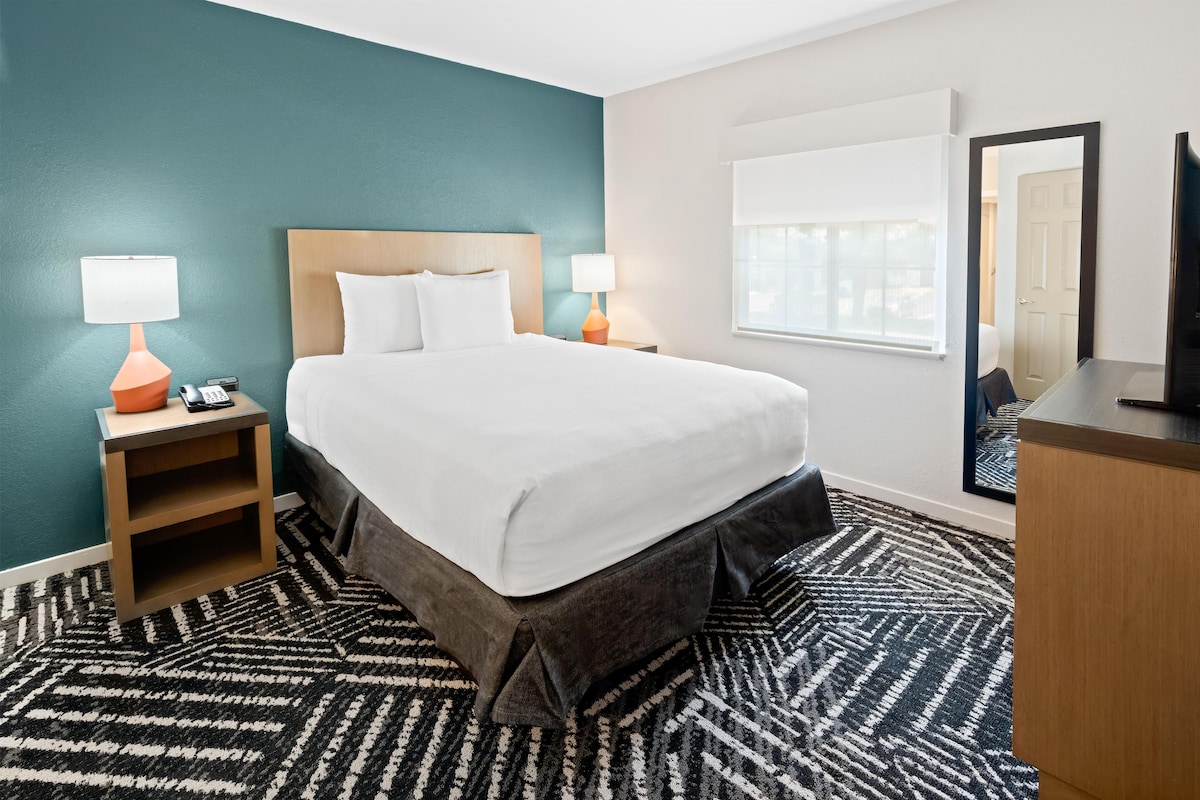 Una habitación de hotel con cama, dos mesitas de noche con lámparas, un espejo y una ventana.