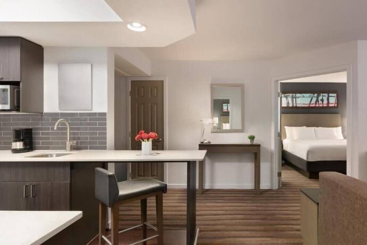 A modern hotel room with a kitchenette, a bed, a TV, and a seating area.