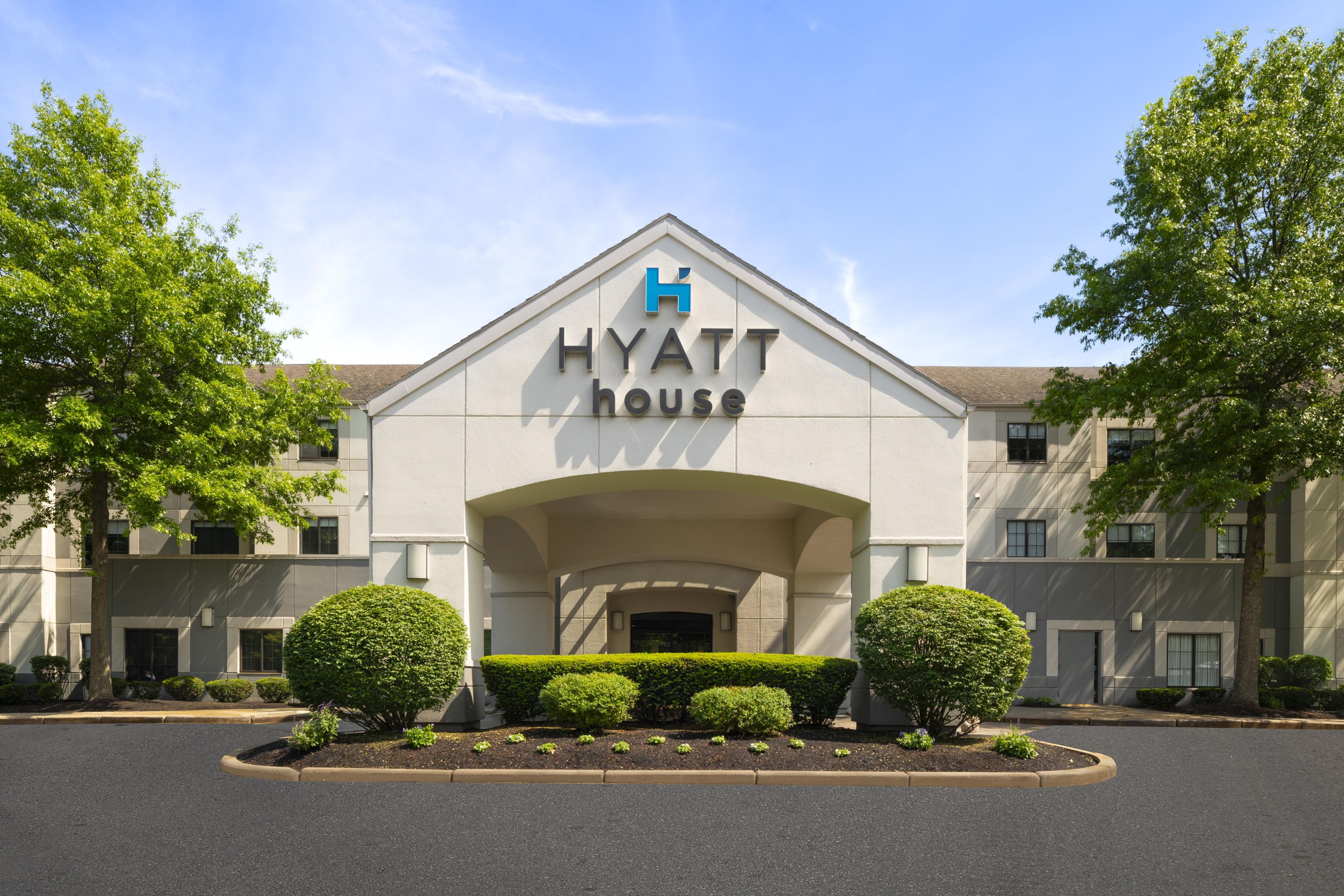A Hyatt hotel entrance with a prominent 'Hyatt House' sign, a landscaped front yard with trimmed bushes, and a clear sky.