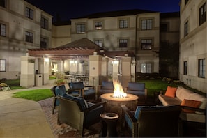 Un patio exterior con mobiliario de mimbre, una chimenea central y una zona de estar cubierta con mesas y sillas.