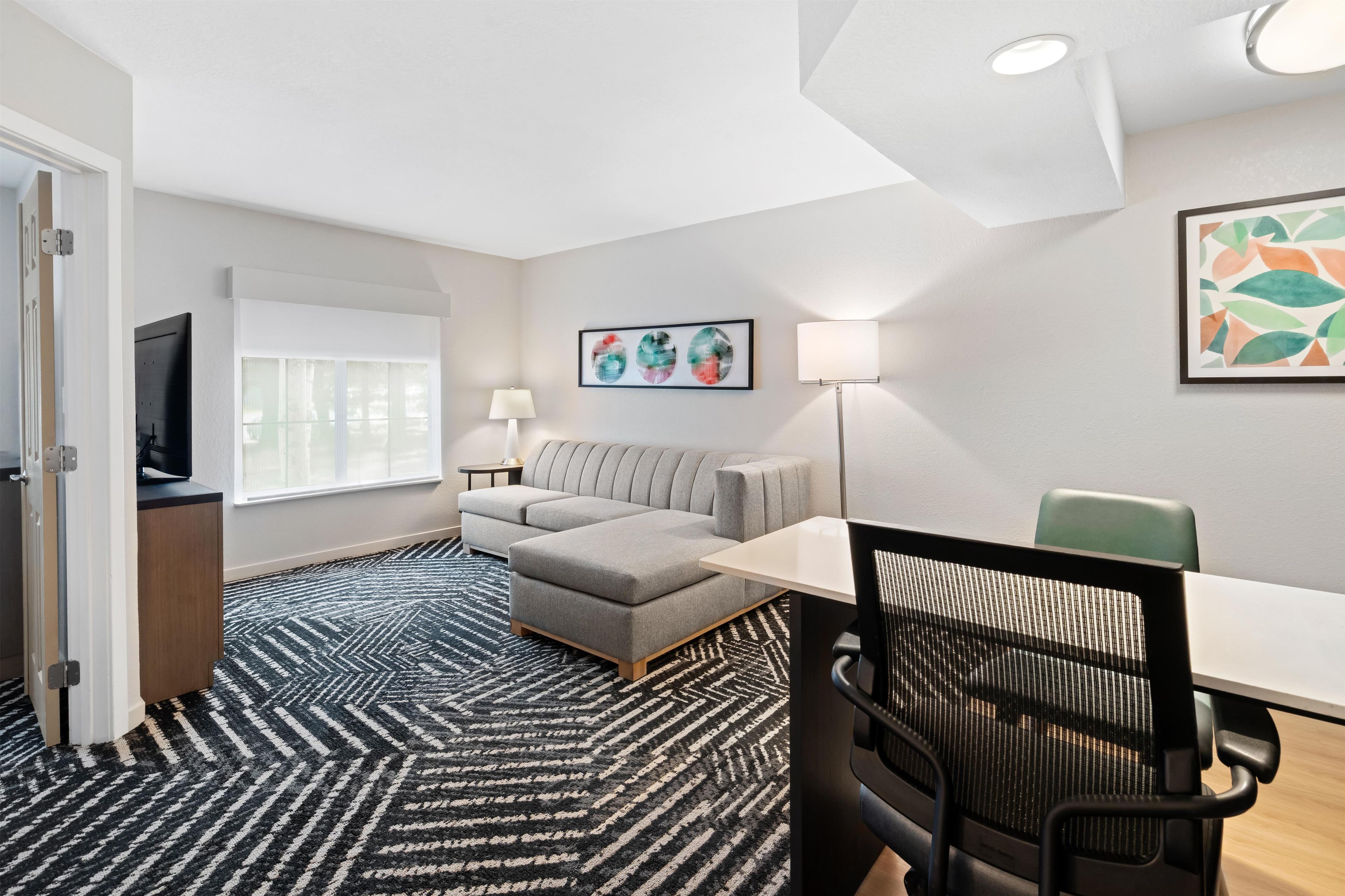 Moderni hotellihuone, jossa on sohva, työpöytä, tuoli, televisio ja taideteoksia seinillä.