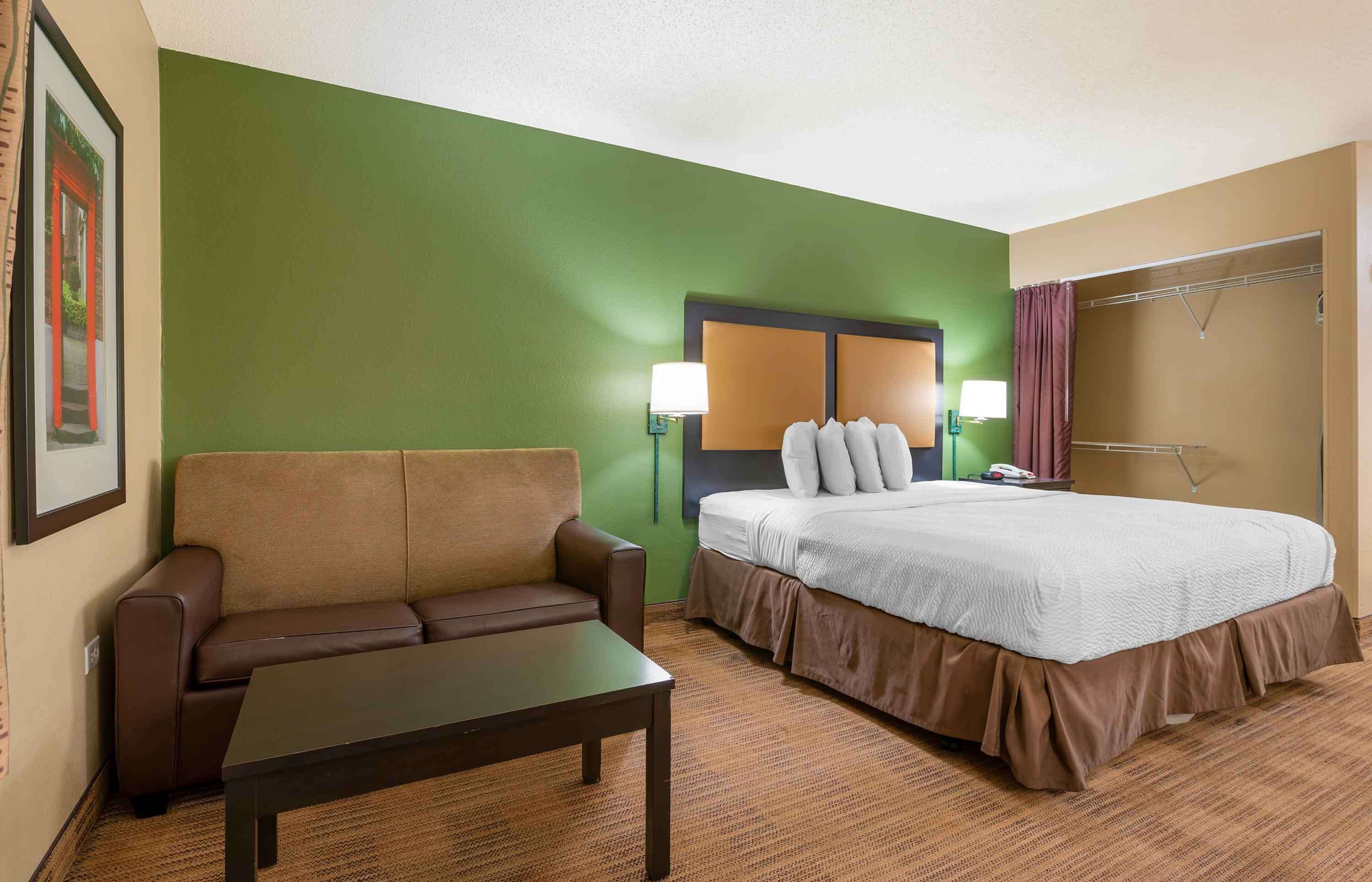 A hotel room with a green accent wall, a brown sofa, a bed with white bedding, and two wall-mounted lamps.