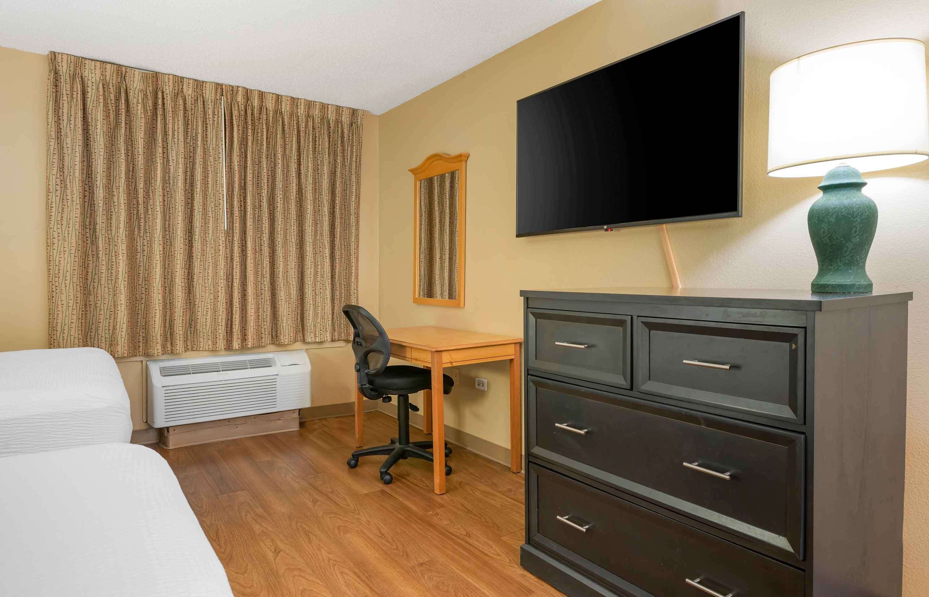 Una habitación de hotel con cama, escritorio con silla, televisor, lámpara y cómoda.