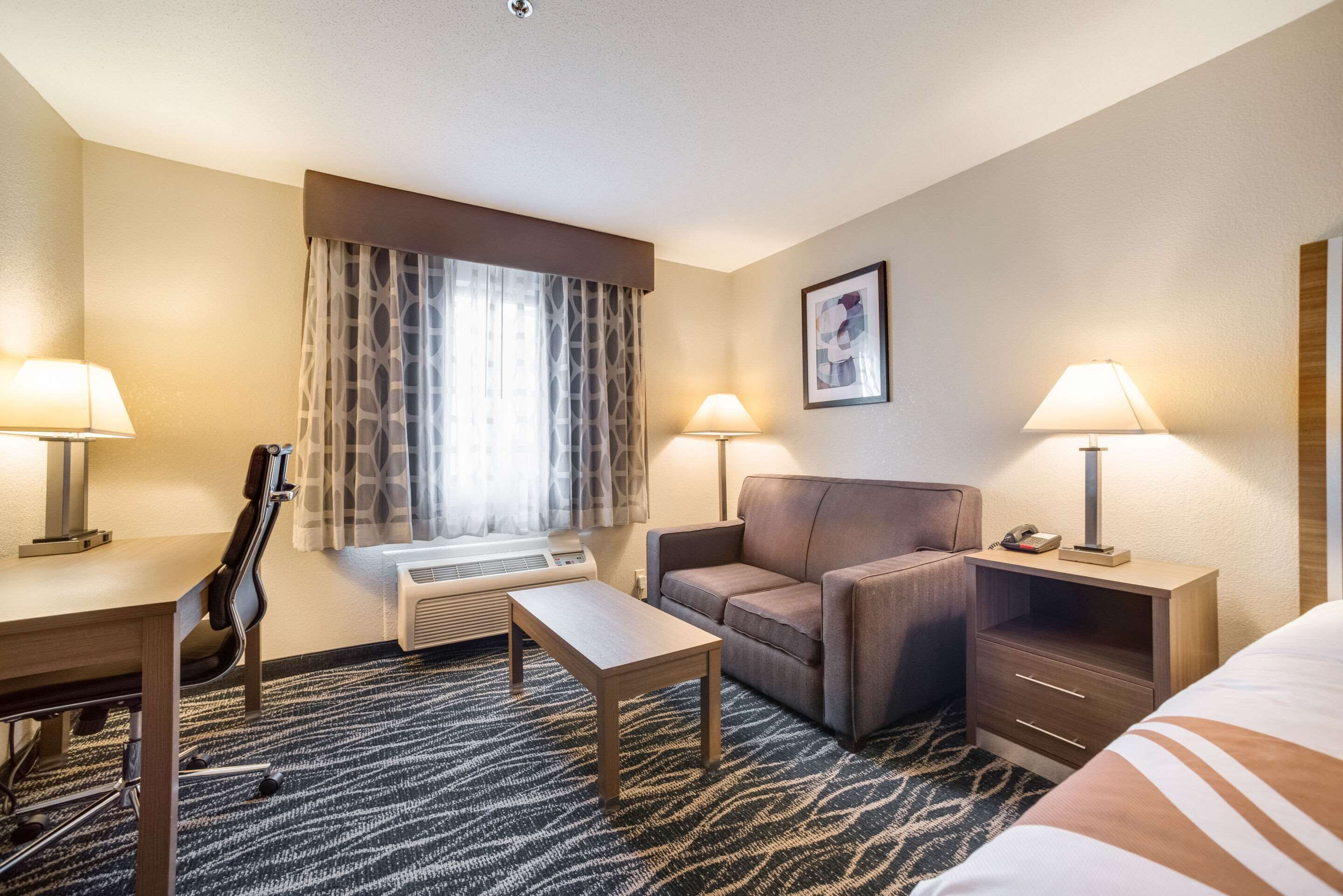 A hotel room with a desk, a chair, a sofa, a small table, a lamp, a heater, and a window with curtains.
