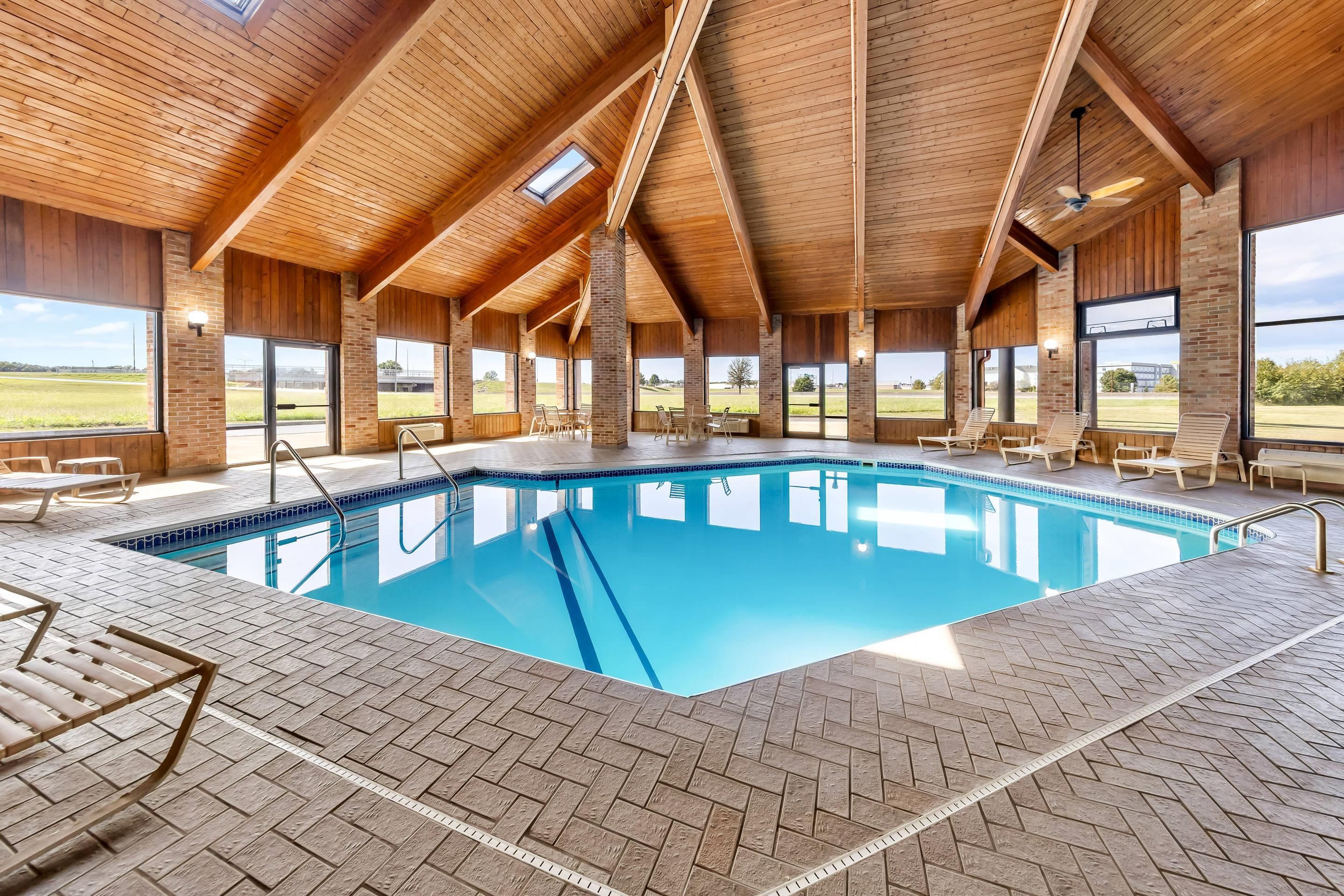 An indoor swimming pool with a tiled floor, wooden ceiling beams, and large windows offering views of the outdoors.