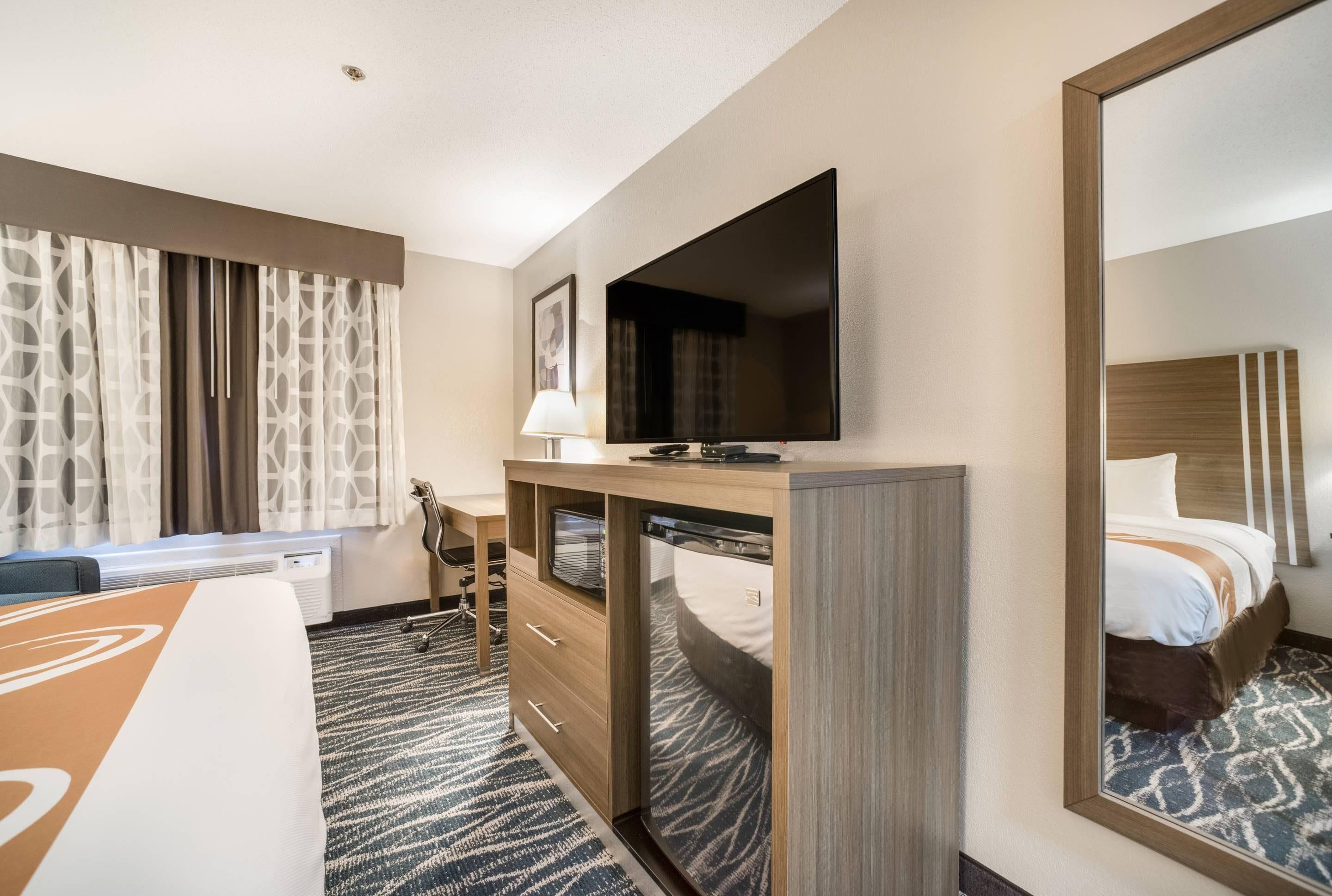 Une chambre d’hôtel comprenant un lit, un bureau avec un téléviseur à écran plat, une chaise, une lampe de bureau et un grand miroir.