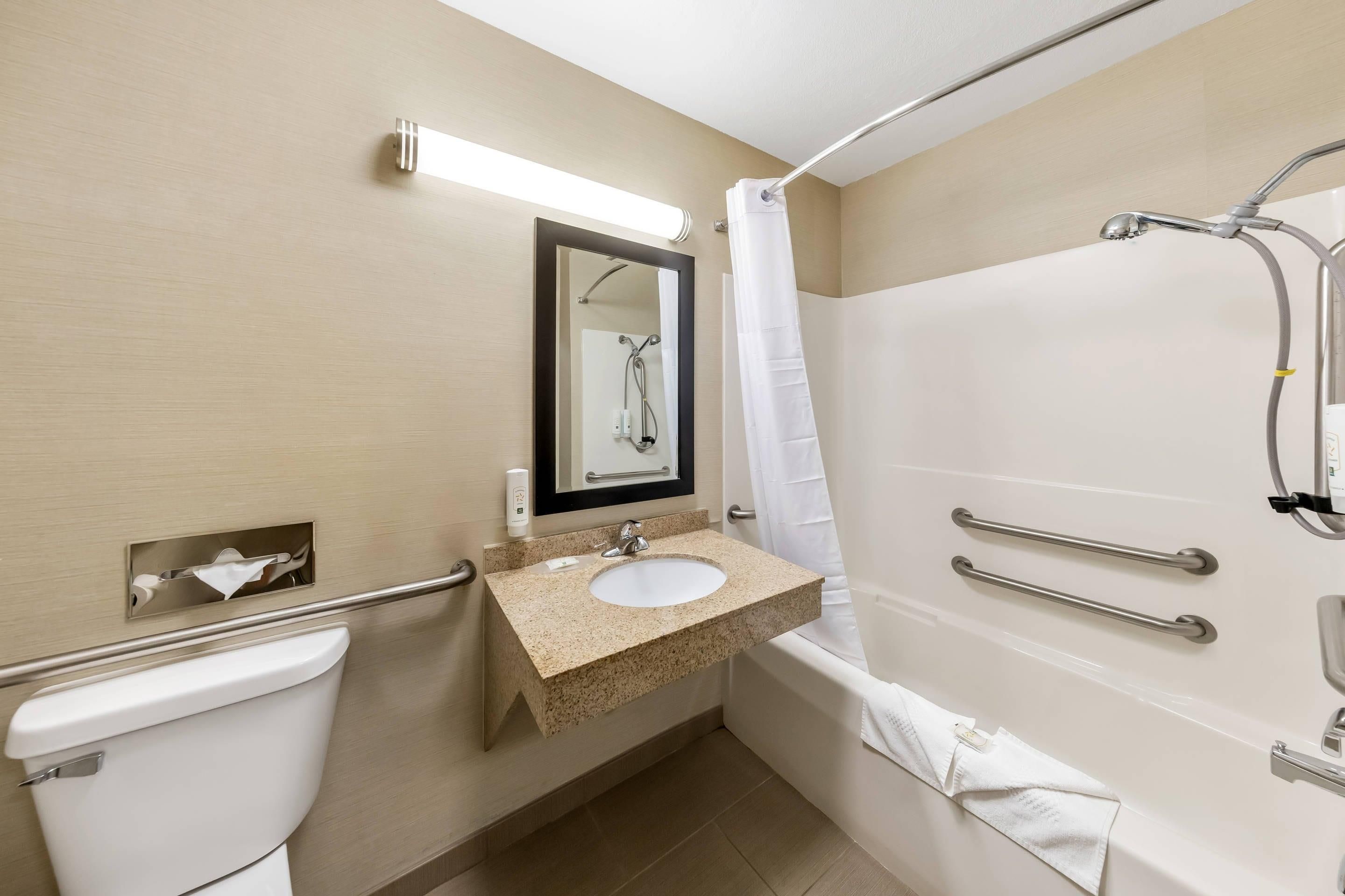 A bathroom with a toilet, sink, mirror, shower, and grab bars.