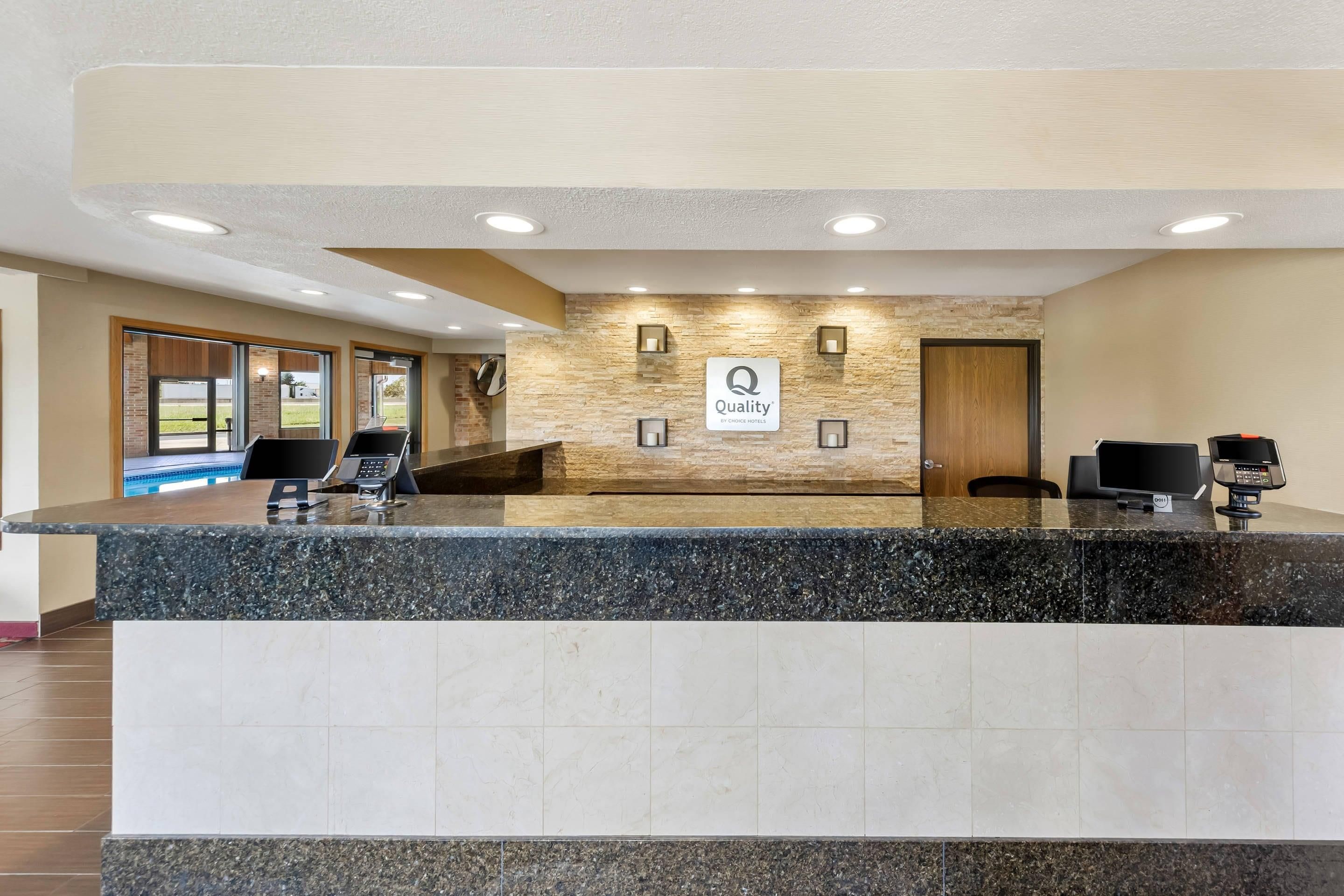 Zona de recepción de hotel con pared de piedra, un cartel que dice 'Quality by Choice Hotels' y un mostrador de recepción con una computadora y un teléfono.