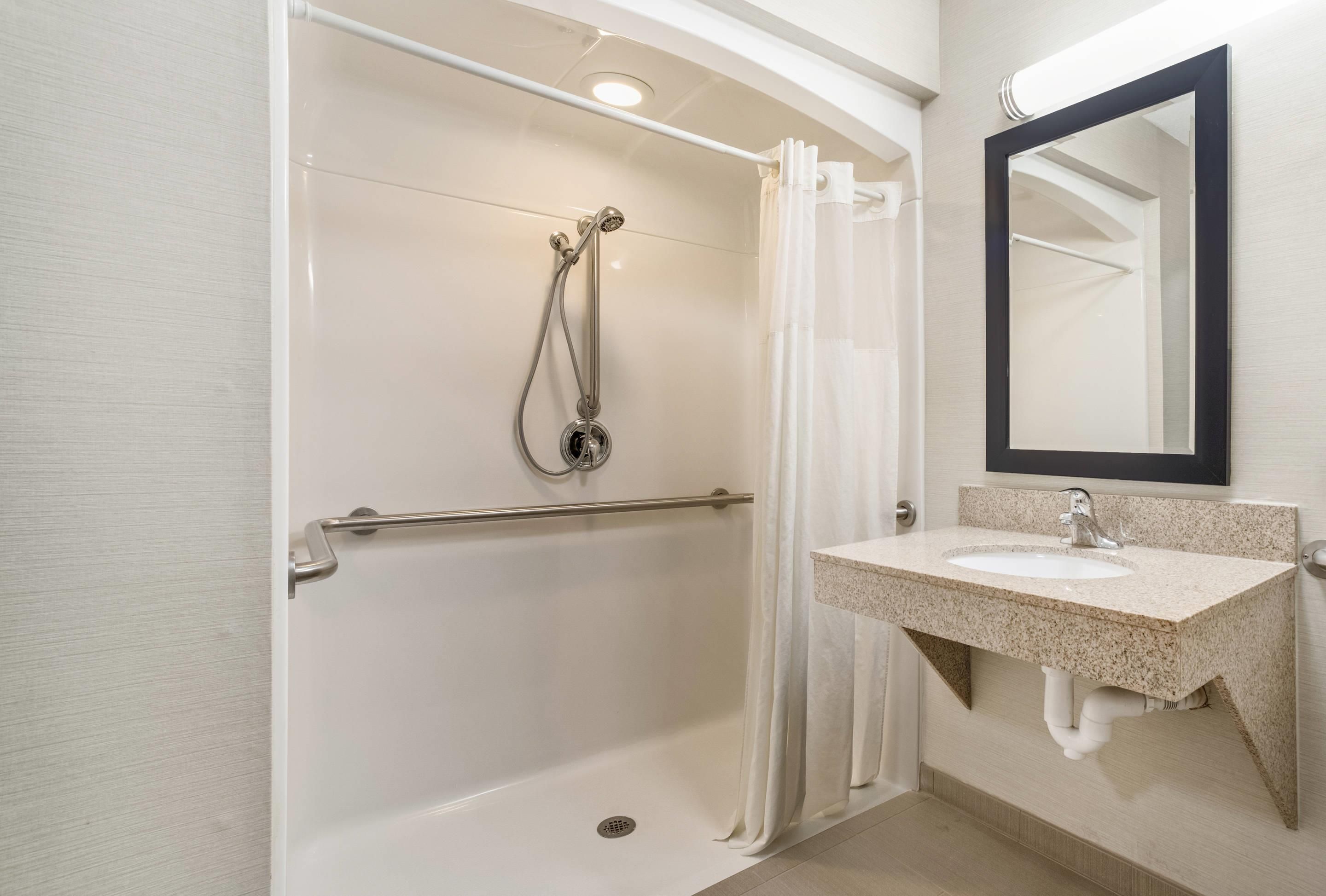 A bathroom with a shower, a handrail, a sink with a mirror, and a towel rack.