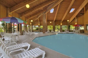 An indoor swimming pool with white lounge chairs and umbrellas, wooden beams, and large windows. - Baymont by Wyndham Muskegon (Muskegon)