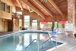 An indoor swimming pool with a wooden deck, lounge chairs, and a poolside table area. - Baymont by Wyndham Muskegon (Muskegon)