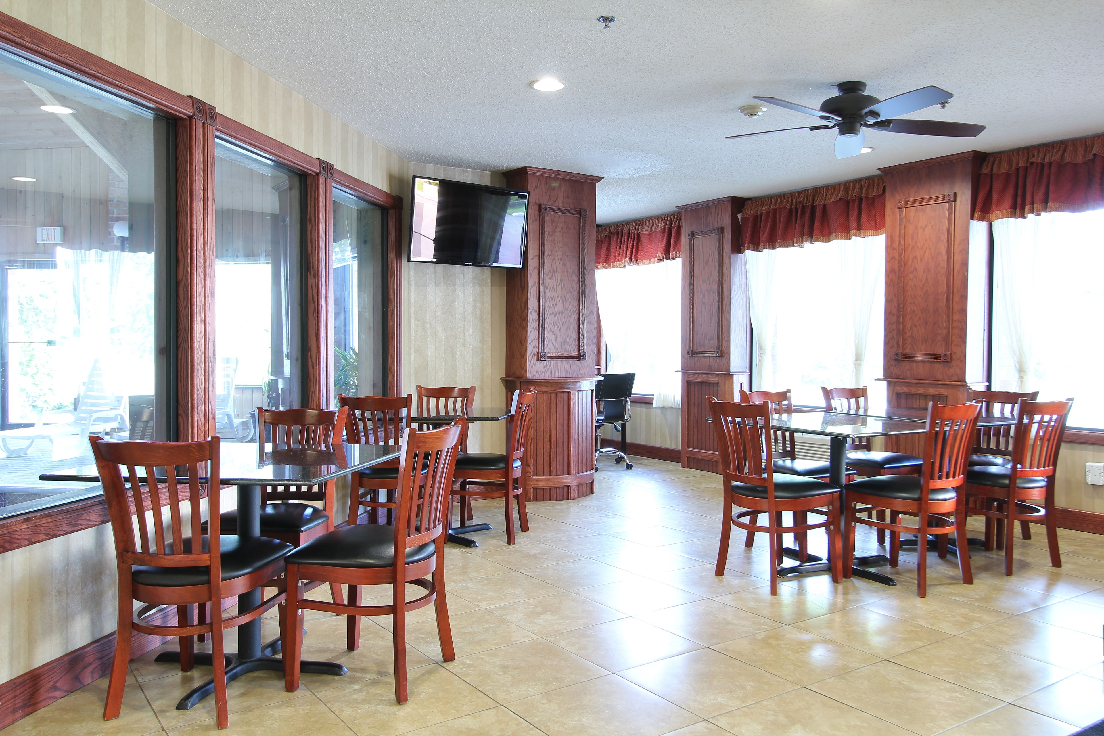 Un comedor con sillas y mesas de madera, un televisor montado en la pared y ventanas grandes con cortinas.