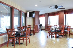 Un comedor con sillas y mesas de madera, un televisor montado en la pared y ventanas grandes con cortinas.