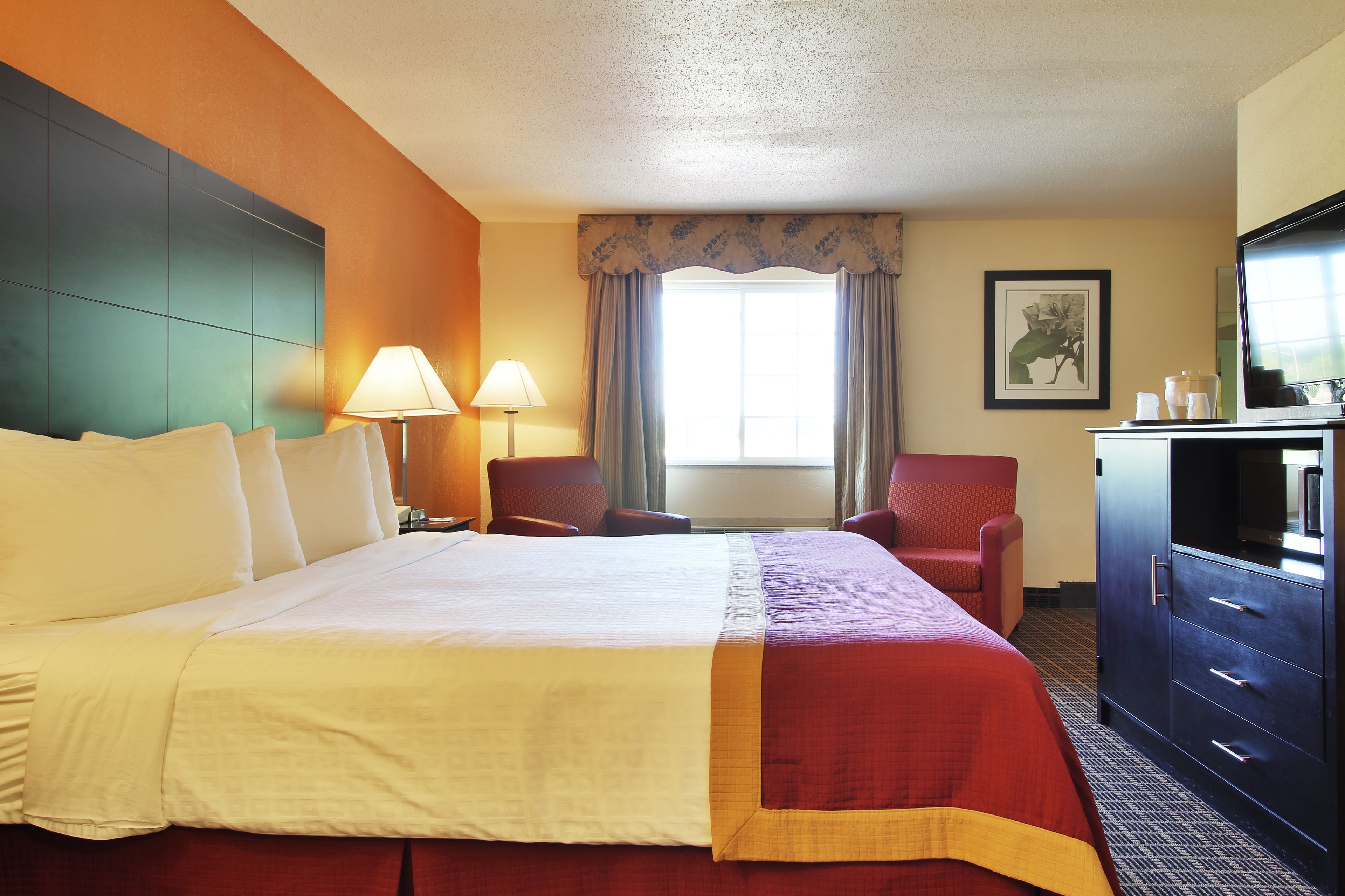 A hotel room with a large bed, two chairs, a desk with a TV, and a window with curtains.