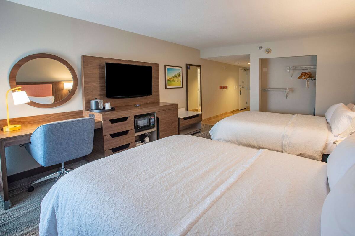 A hotel room with two beds, a desk with a chair, a TV, a mirror, and a wardrobe.