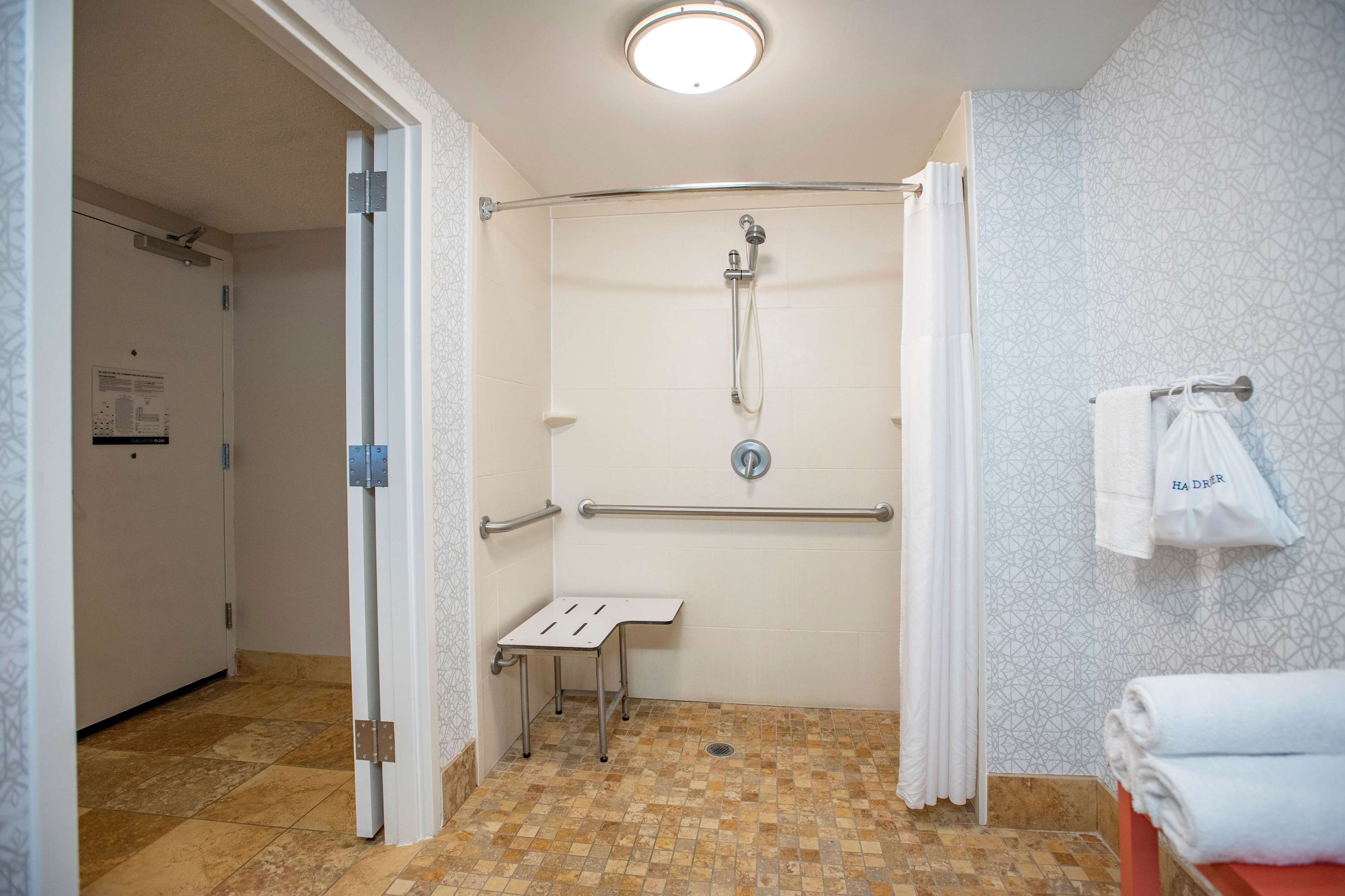 A wheelchair-accessible shower with a handrail, a small table, and a towel rack with a towel labeled "HA DRER".