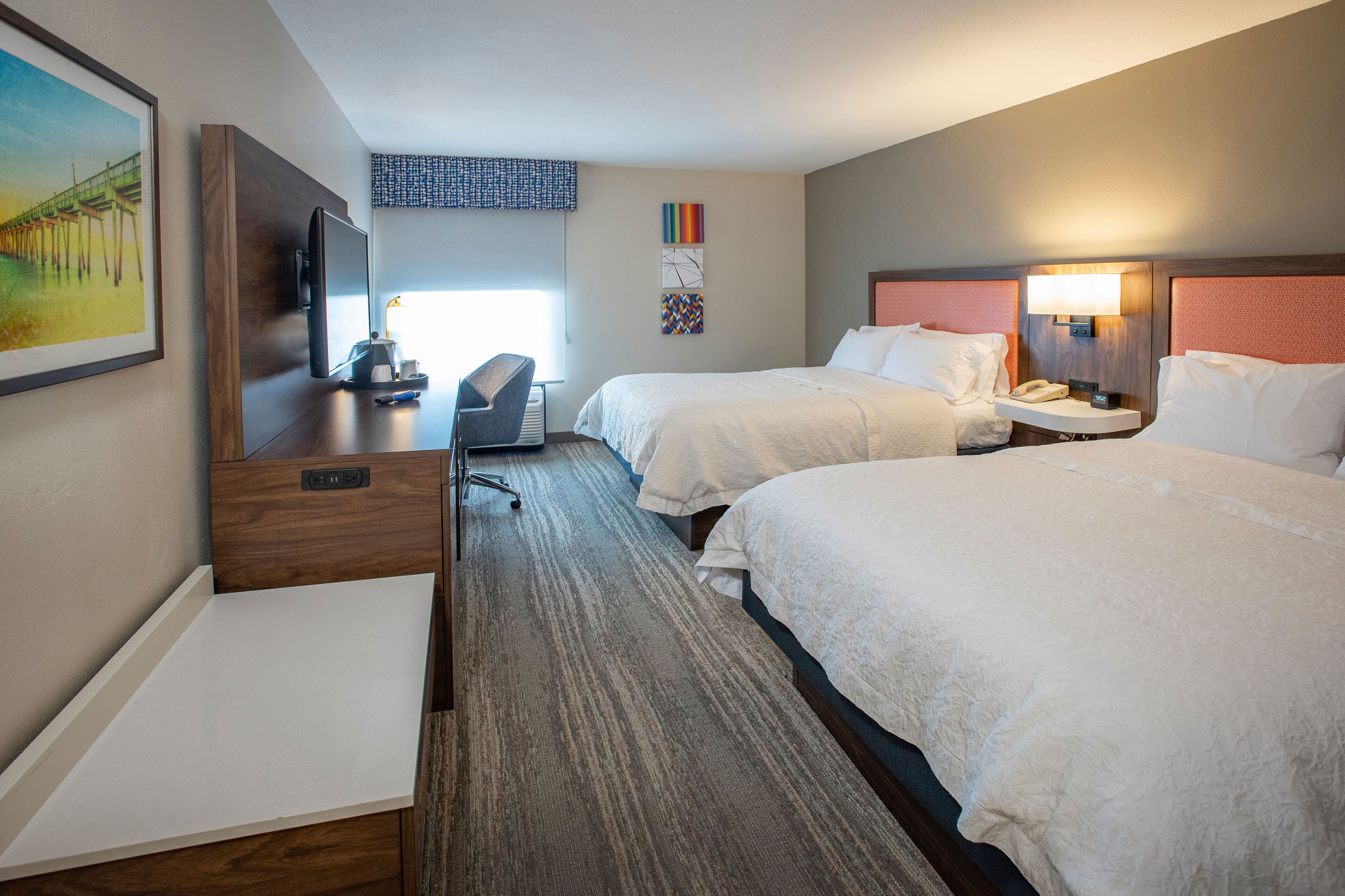 A hotel room with two beds, a desk, a chair, a TV, and a painting on the wall.