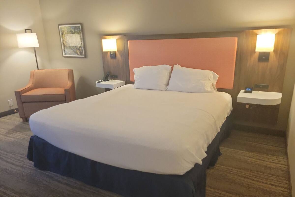A hotel room with a large bed, a chair, a nightstand, a wall-mounted lamp, and a painting.