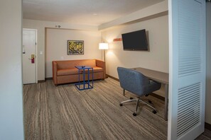 A hotel room with a sofa, a desk, a chair, a lamp, and a flat-screen TV.