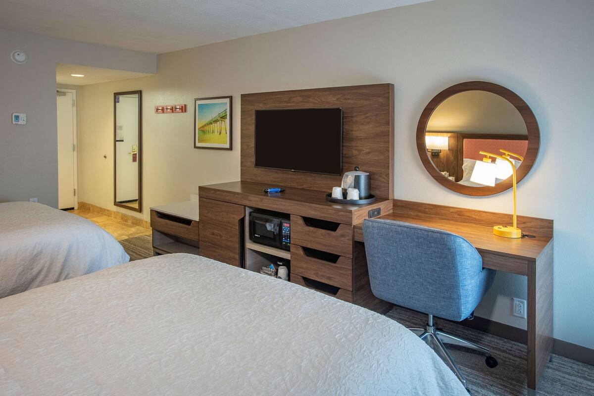 A hotel room with two beds, a desk with a flat-screen TV, a microwave, a coffee maker, and a round mirror.