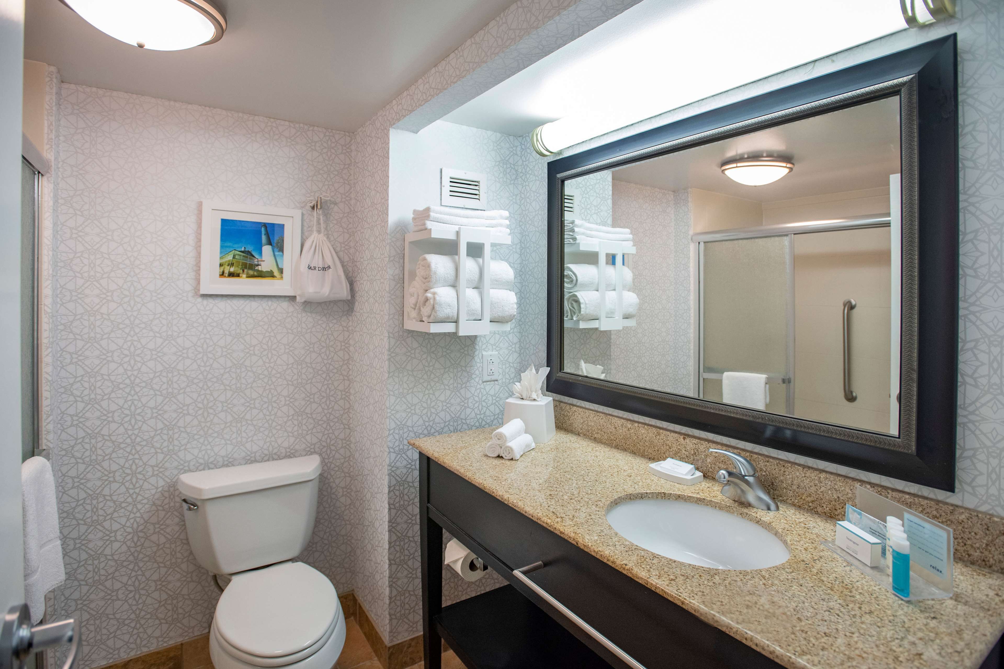 Un baño moderno con inodoro, lavamanos y un espejo grande. Hay toallas y artículos de aseo en la encimera.