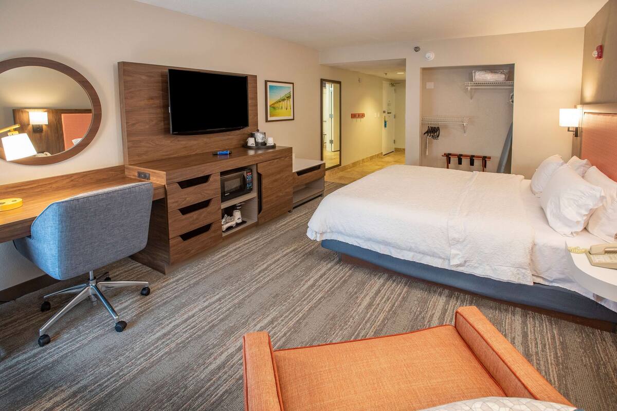 A hotel room with a bed, a desk with a chair, a TV, a mirror, and a door leading to a bathroom.