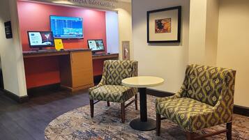 A seating area with patterned chairs and a small table, a desk with monitors displaying flight information, and a framed artwork on the wall.