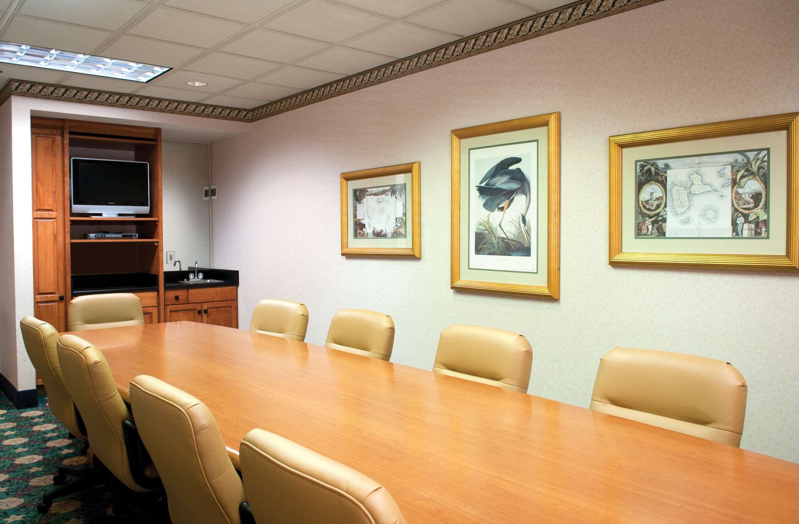A conference room with a long wooden table, six beige chairs, a television on a wooden cabinet, and three framed pictures on the wall.