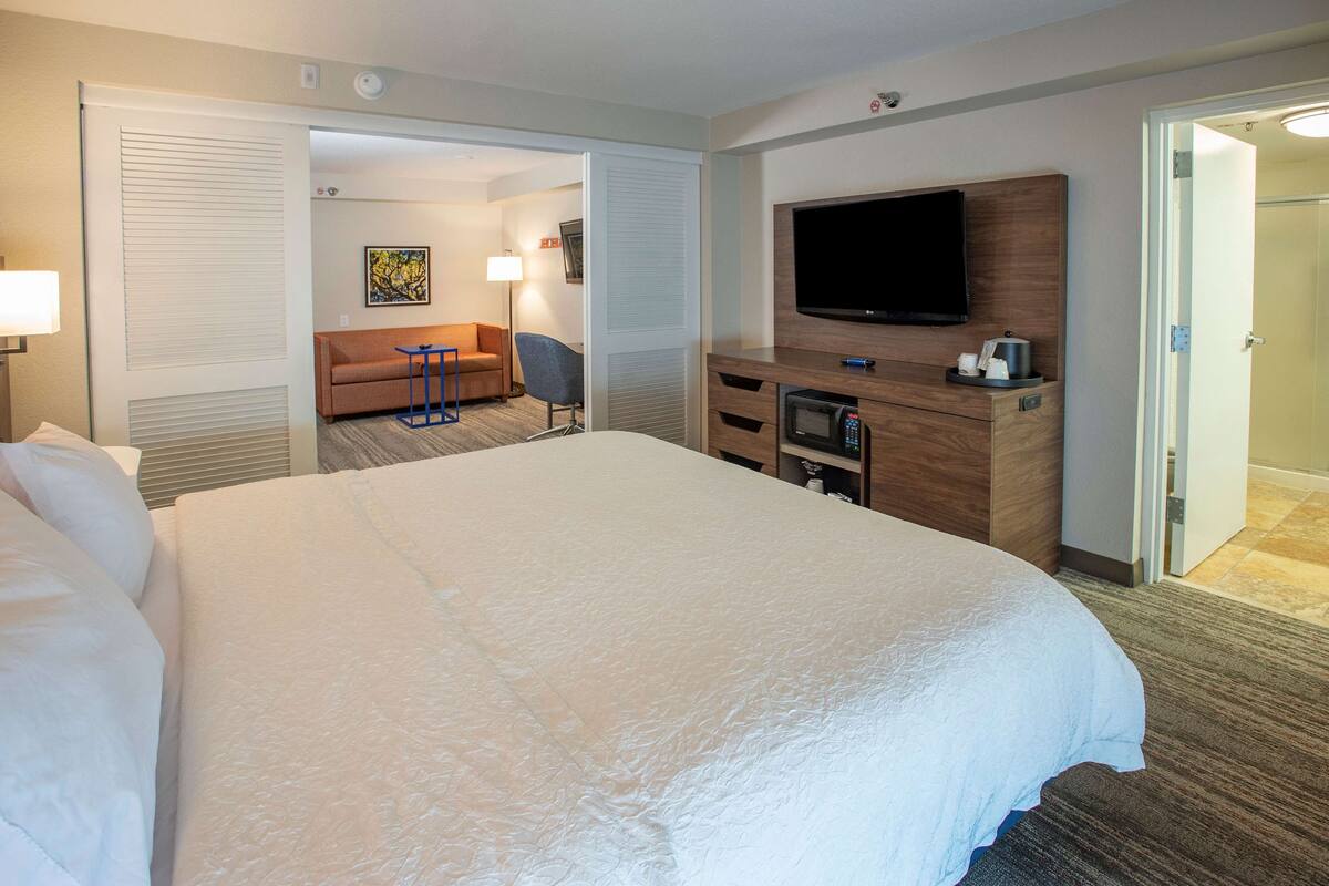 A hotel room with a large bed, a flat-screen TV, a desk with a microwave, and a sofa in the background.