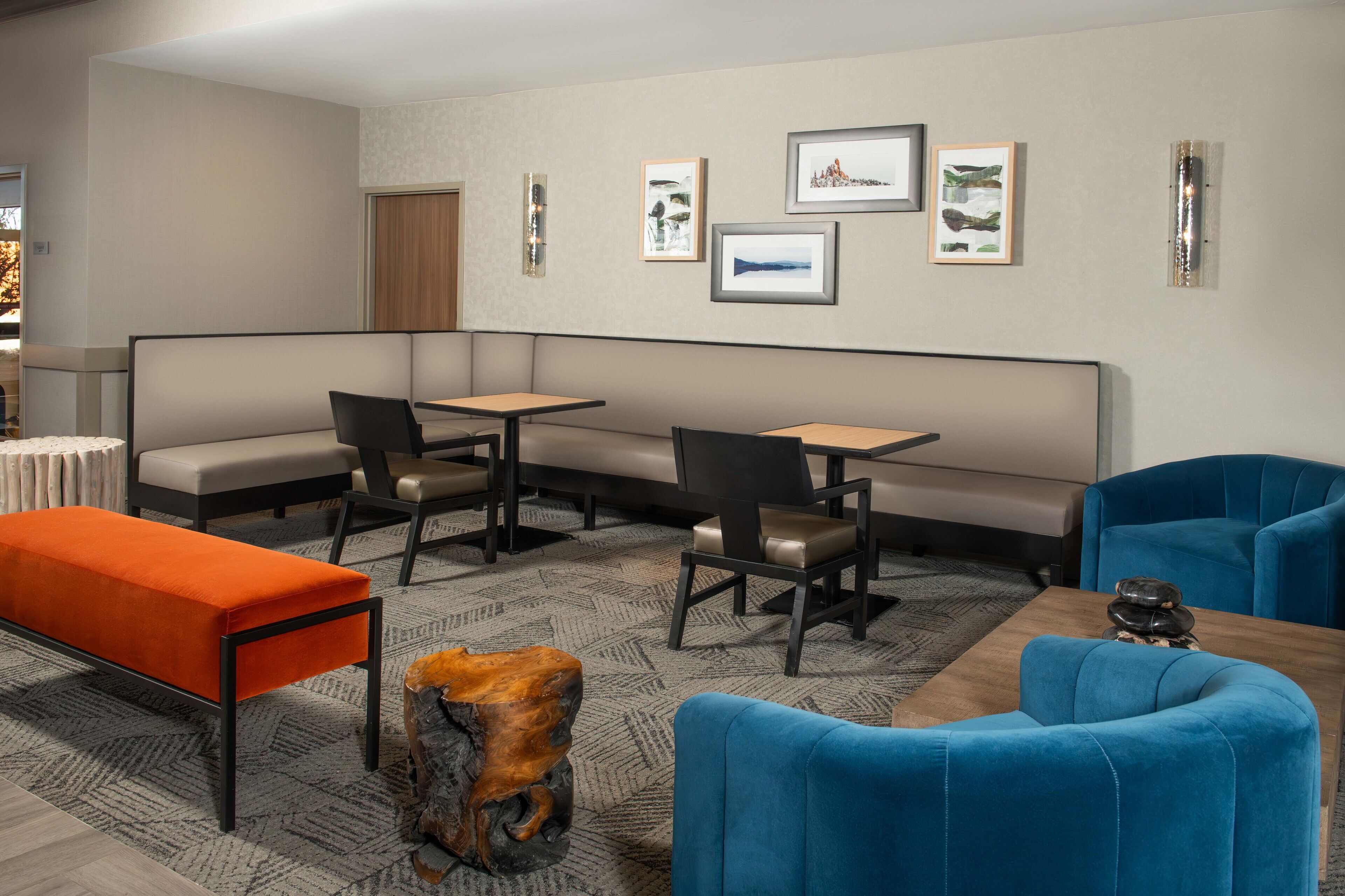 A modern hotel room with a sectional sofa, a dining table, and a small coffee table. There are framed pictures on the wall and a unique wooden stool.