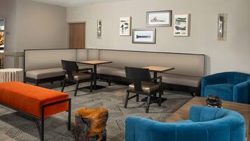Ein modernes Hotelzimmer mit Ecksofa, Esstisch und kleinem Couchtisch. An der Wand hängen gerahmte Bilder und ein außergewöhnlicher Holzschemel.