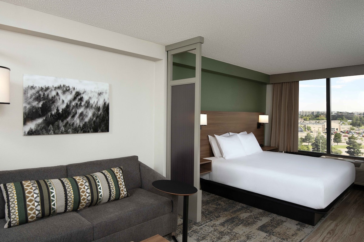 A hotel room with a bed, a sofa, a small table, a painting, and a view of the city.