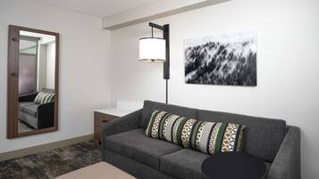 A modern living room with a grey sofa, patterned cushions, a round side table, a floor lamp, a framed abstract painting, and a mirror reflecting the room.
