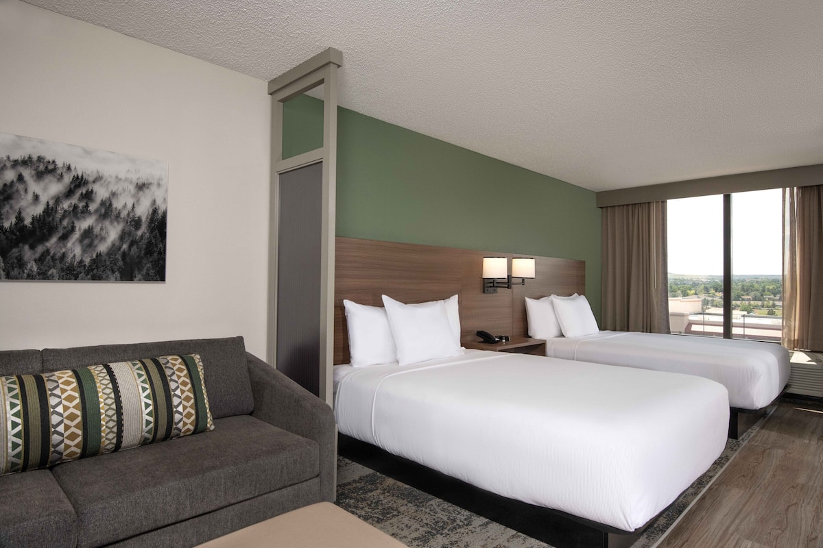 A hotel room with two beds, a sofa, a painting, and a large window with curtains.